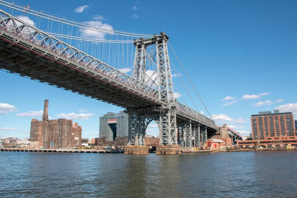 Visiting New York offers iconic sights like the Williamsburg Bridge spanning the East River, set against a blue sky and urban buildings in the background. Discover the city’s charm now!