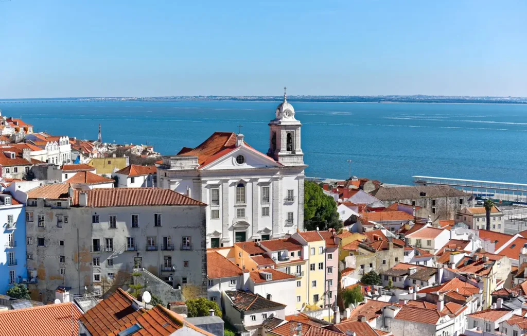 Here's Why visit Lisbon, Portugal. The cityscape with colorful buildings and red tile roofs overlooking the Tagus River.