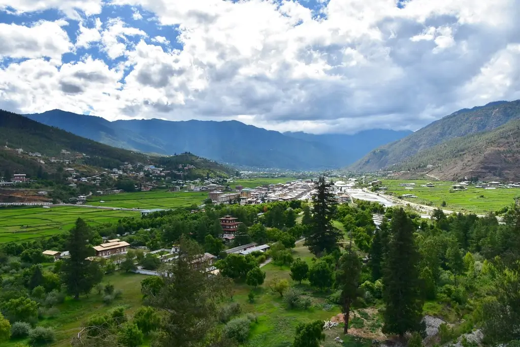 Panoramic view of Paro Valley, Bhutan with rice paddies, traditional pagoda, Himalayan mountains, and forests under blue sky.